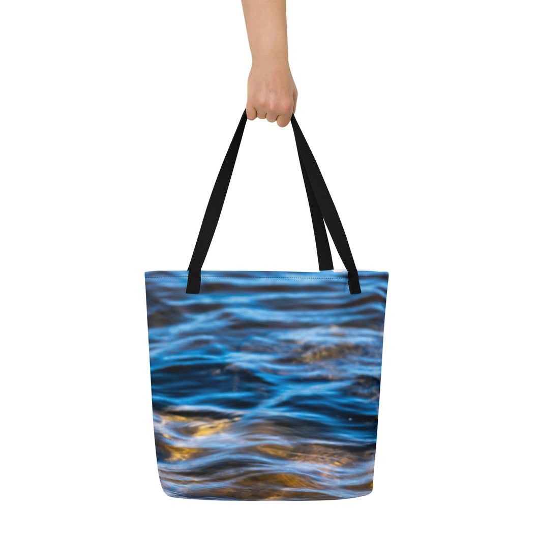 A fully printed tote bag, blue crystal clear water and rocks at the bottom, black beams, carried by one hand. Front side